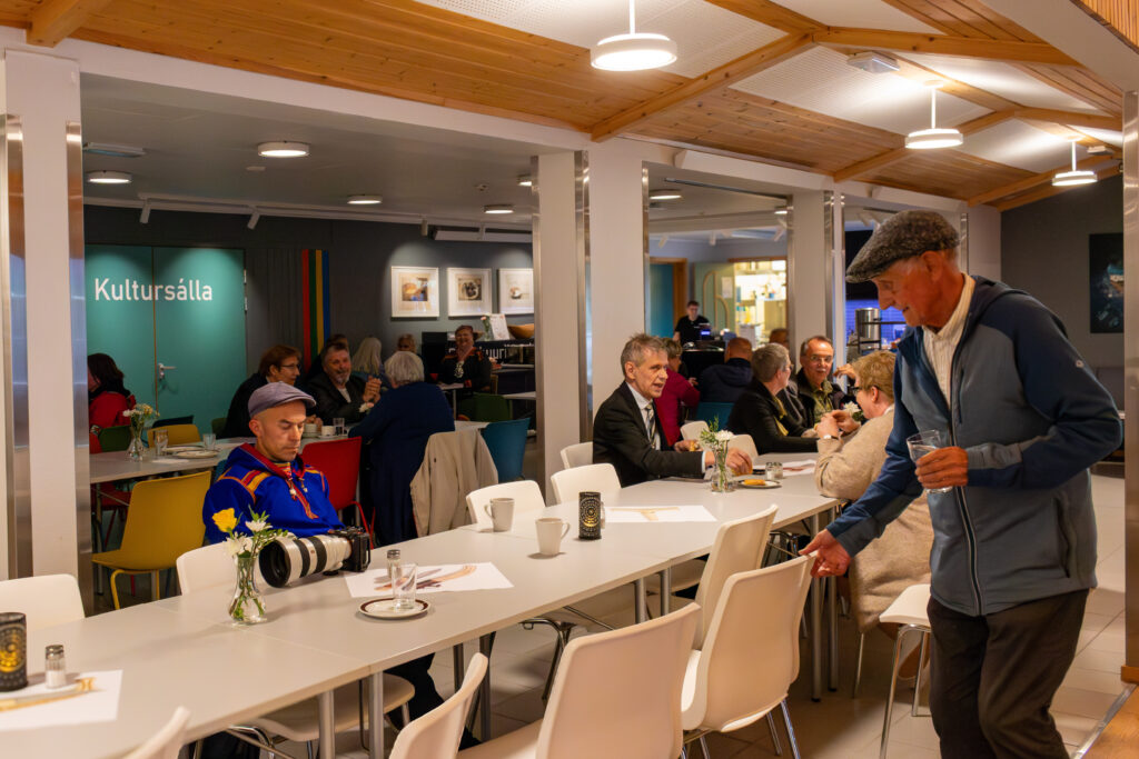 People sit at white tables in a bright community hall, a server pouring drinks on the right.