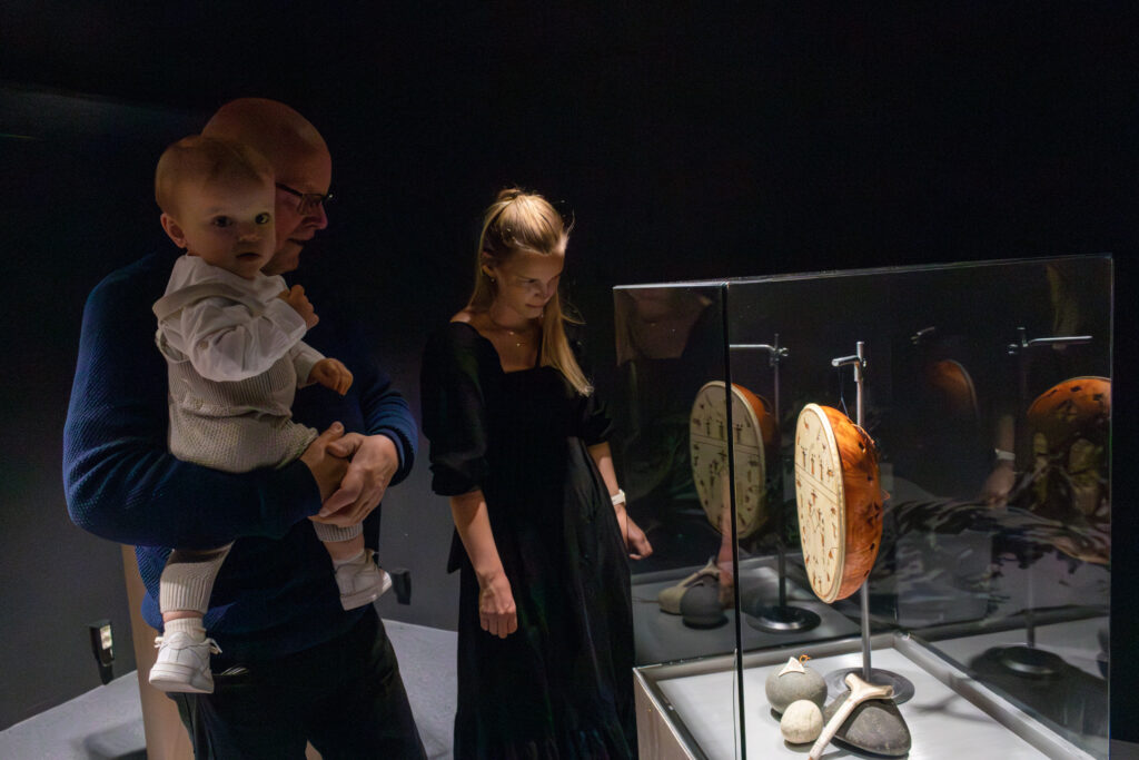 Father holds a baby as a woman and child look at a museum display case with ancient artifacts in dim lighting on display shelves behind glass.