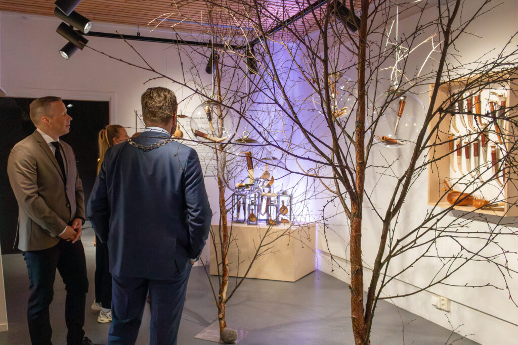 People in a gallery observe a knife exhibit arranged on a wall, with bare tree branches in the foreground creating a natural frame.