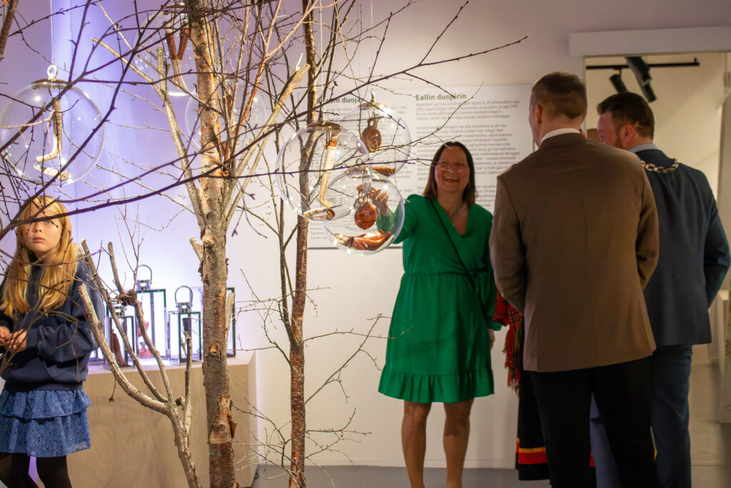 Smiling woman in a bright green dress chats with two men in a gallery, beside a leafless tree sculpture with hanging glass orbs.