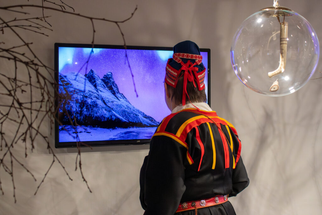 Person in traditional red-and-black clothing stands with back to camera, facing a computer monitor showing a snowy mountain landscape.
