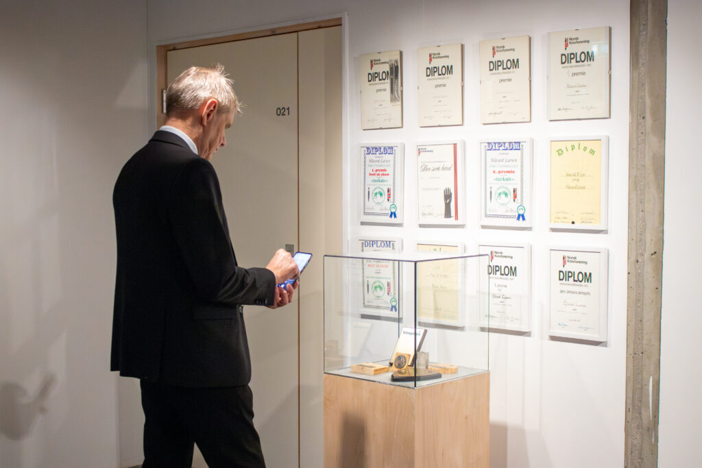 Man in a dark suit studies a wall of framed diplomas in a gallery, holding a phone in his hands at his side.