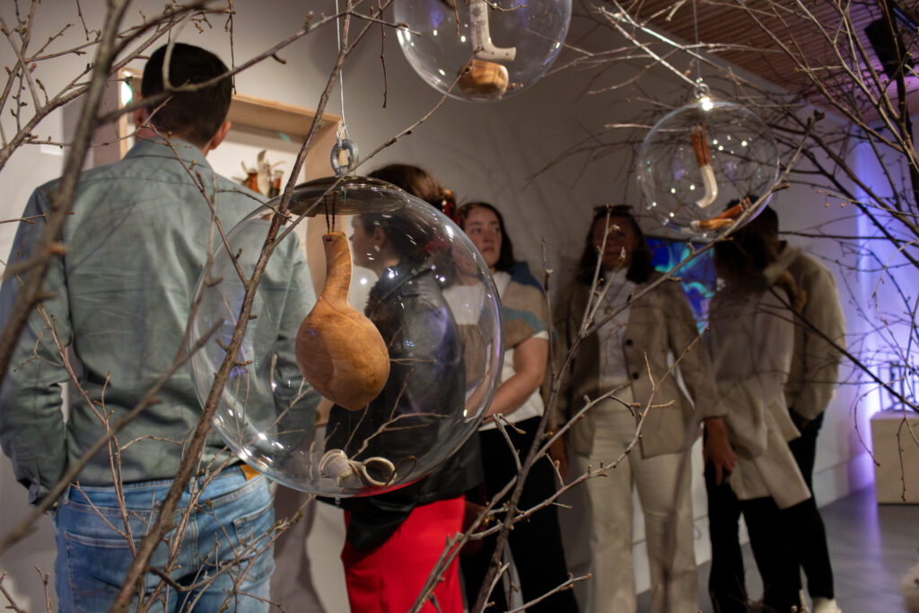 Visitors observe an art installation: bare branches with large clear glass orbs containing wooden pieces hanging among them in a gallery setting.