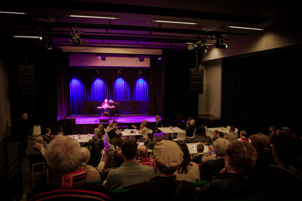 Performer playing keyboard on stage with blue curtains and purple lighting, audience seated at tables in a dim venue, watching.