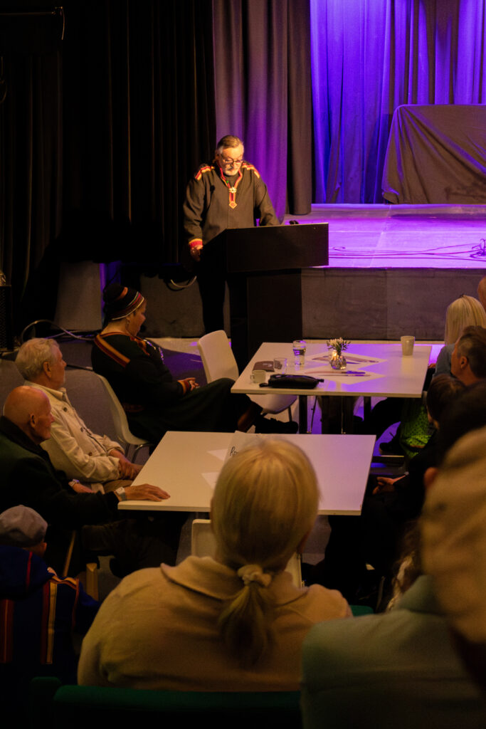 Speaker at a lectern delivering a talk on a purple-lit stage to an attentive audience.