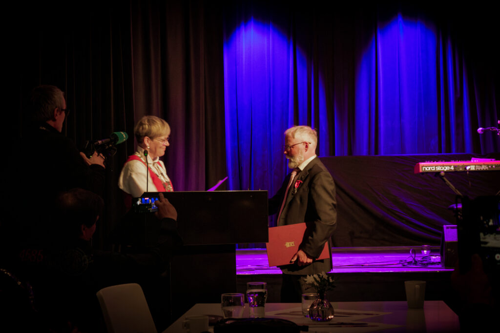 Two people on a dimly lit stage: a man in a suit receiving a red folder from a woman at a podium while a photographer films the moment with a green mic held nearby. Purple stage curtains in the background.