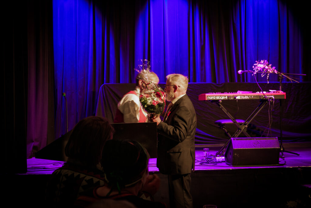 Man in a suit accepts a bouquet on a dimly lit stage with blue curtains, a piano keyboard to the right and a microphone nearby.