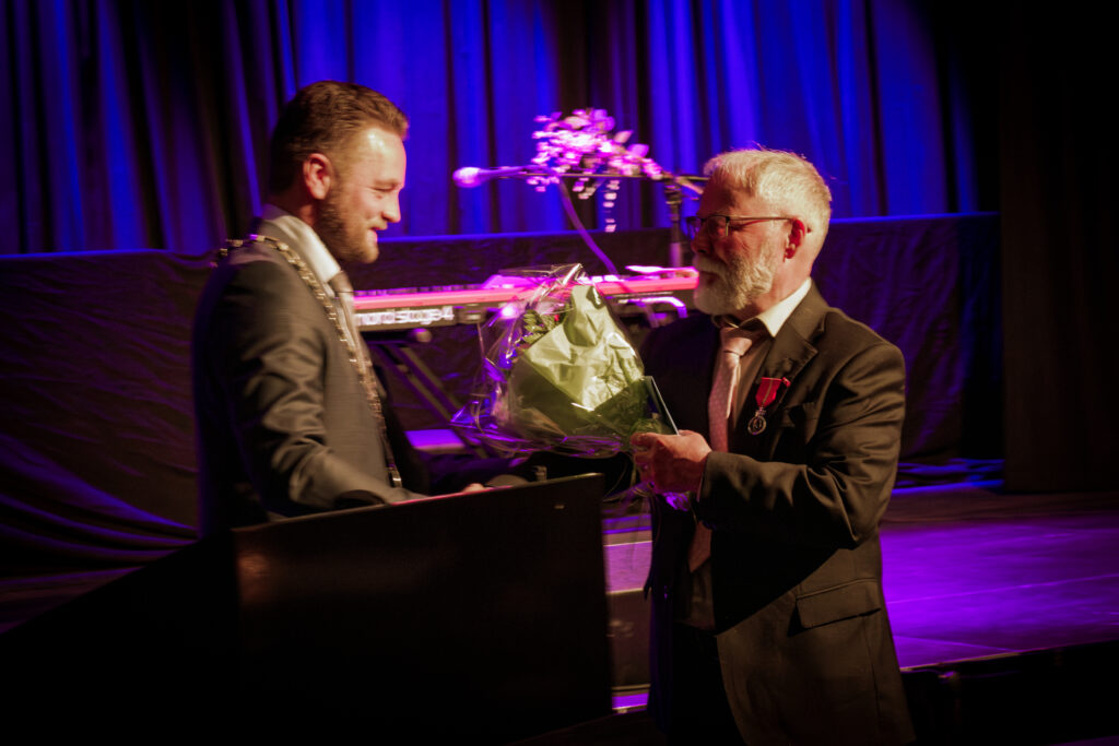 Two men in suits on a stage exchanging a bouquet, with purple stage lighting and a keyboard in the background.