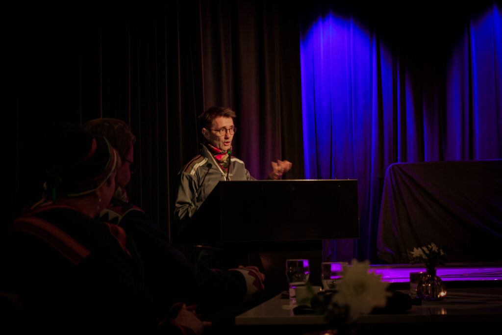 Performer at a piano on a dim stage with blue lighting, addressing an audience in the foreground.