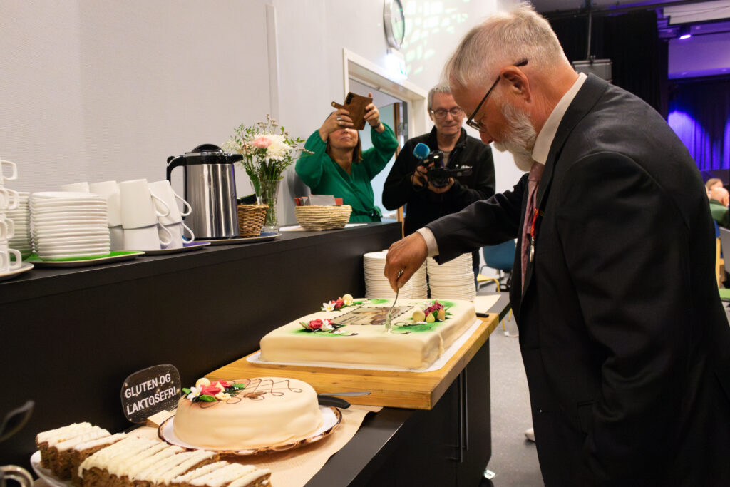 Man in a suit slices a decorated cake at a buffet while photographers capture the moment.