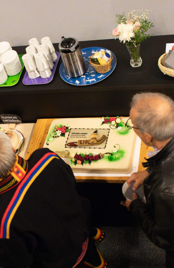 People gathered around a large decorated cake on a table, with visible floral icing and green accents, while others prepare to serve slices.