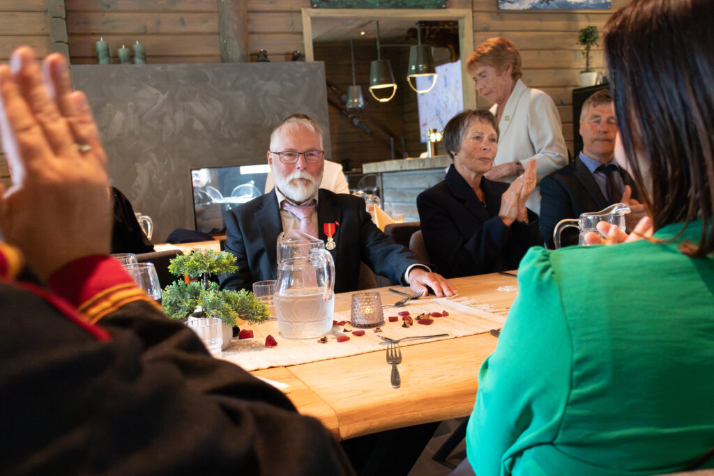 Group of people seated around a wooden table at a celebration, applauding as they converse; a water pitcher and small centerpiece on the table.