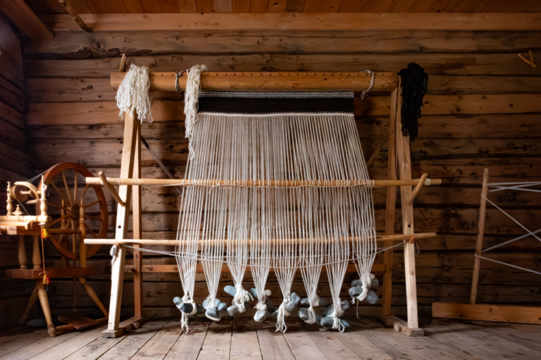 Wooden weaving loom set up in a rustic cabin, with white warp threads hanging and wool at the loom’s heddles on the floor.