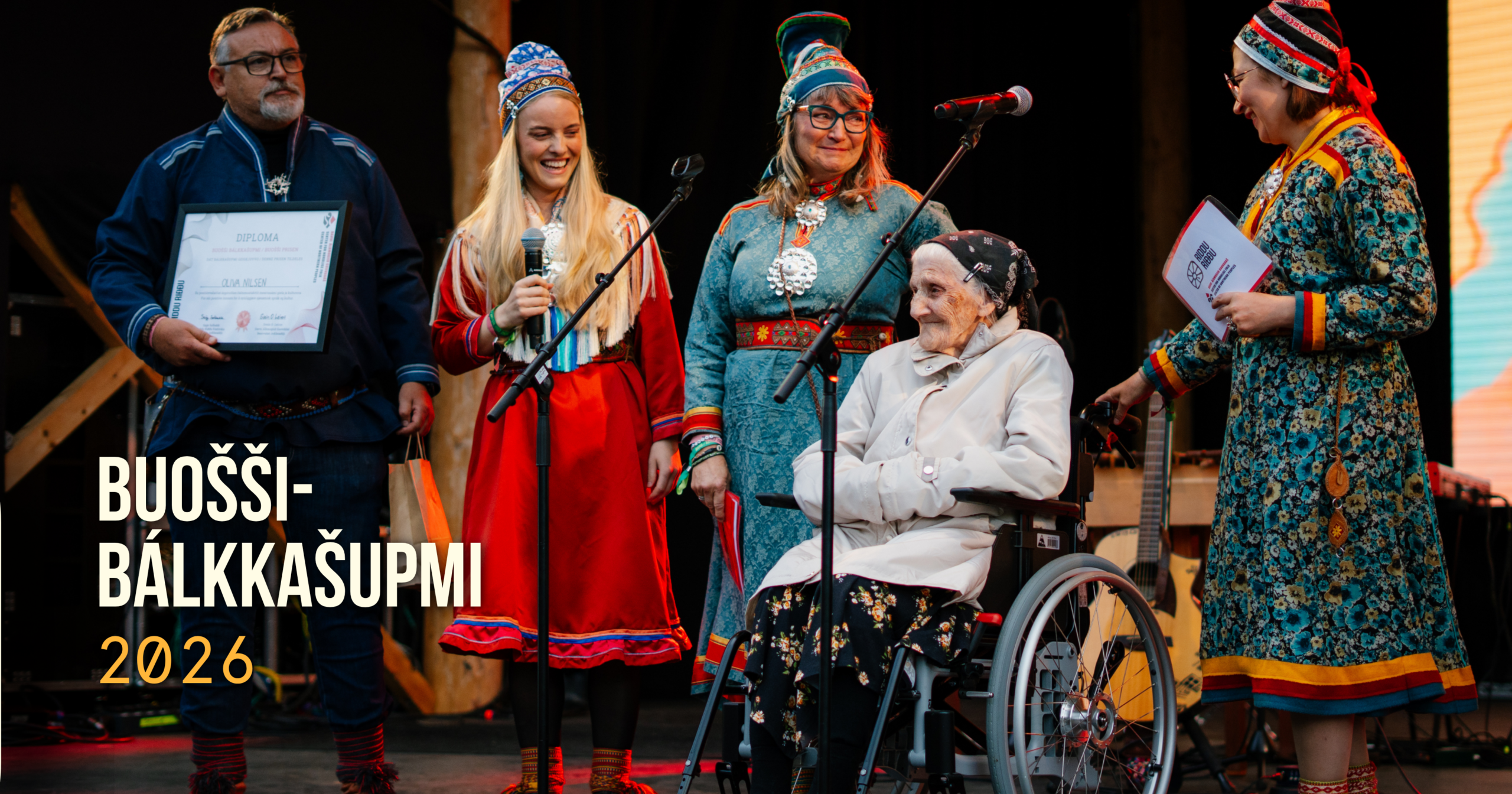 Group of people in colorful traditional outfits on a stage; a man holds a framed diploma, others smile, one woman in a wheelchair sits center-right, microphones nearby.