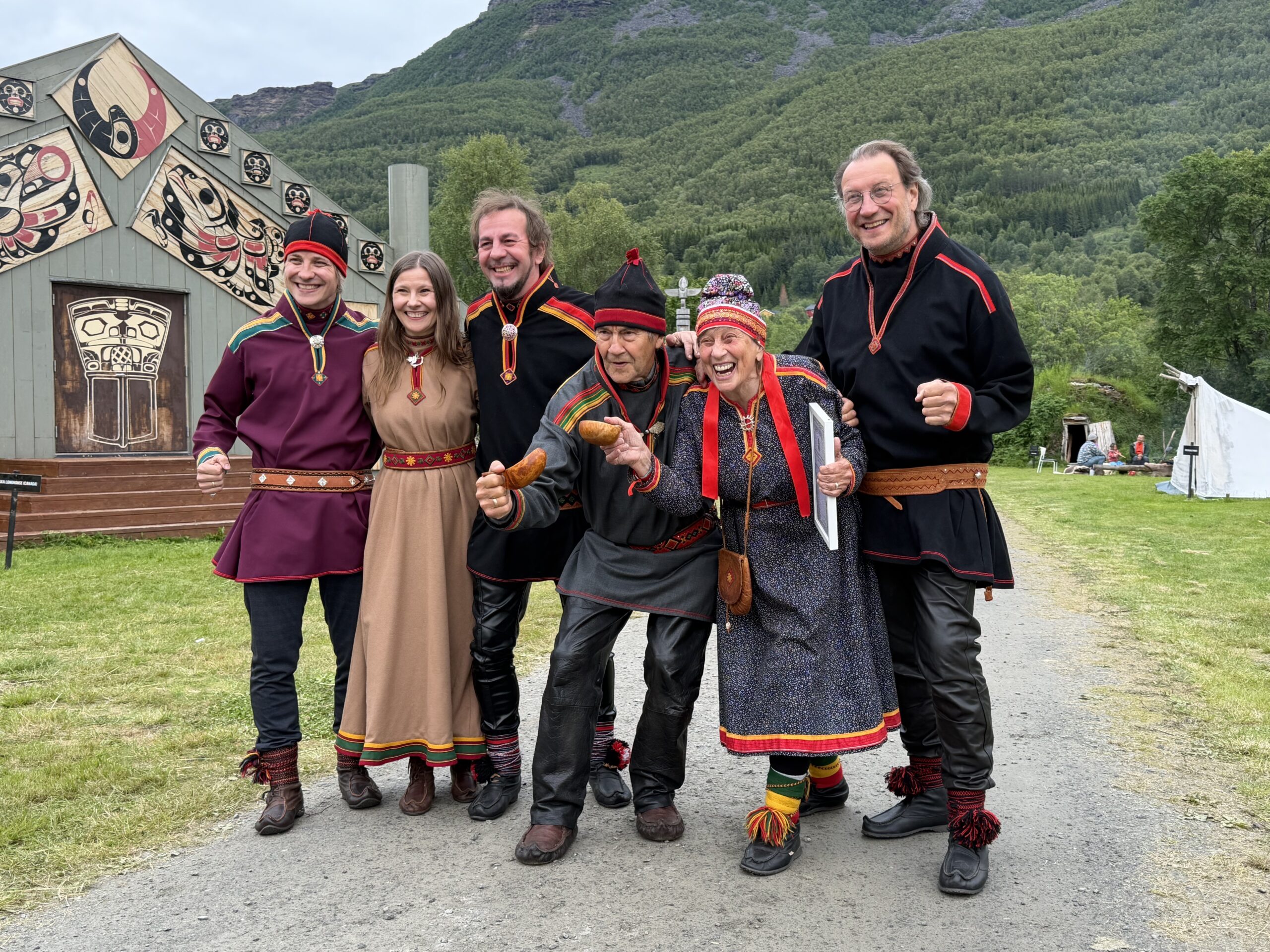 Six people in colorful traditional costumes pose together on a path, with a wooden building and green hills in the background.