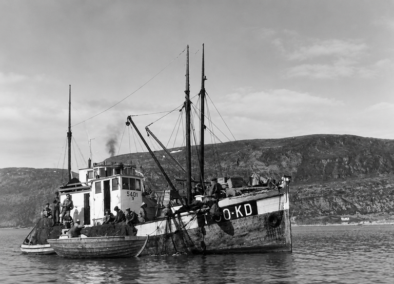 Old fishing trawler with people aboard, anchored near a small rowboat on calm water with rocky hills in the background (black and white).