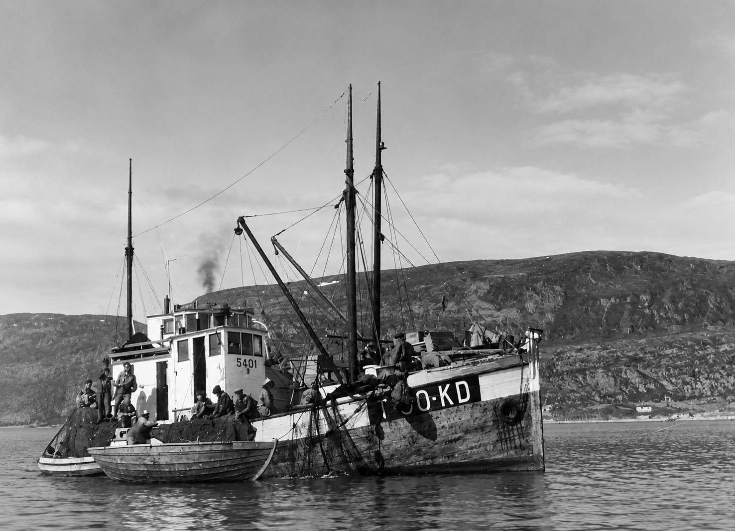 Old fishing trawler with people aboard, anchored near a small rowboat on calm water with rocky hills in the background (black and white).