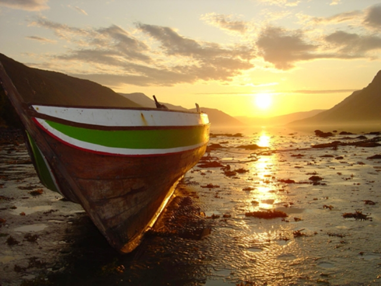 Wooden boat with a green and white stripe rests on a wet, rocky shore at sunset, water reflecting the golden sky.